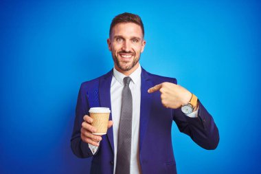 Genç yakışıklı bir iş adamı, mavi izole bir arka planda kağıt bardakta kahve içerken kendini gösteren sürpriz bir yüzle.