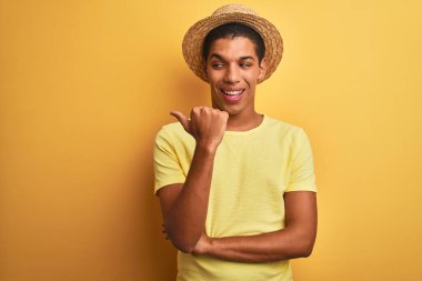 Genç yakışıklı Arap adam tişört giyiyor ve yaz şapkası takıyor izole edilmiş sarı arka planda gülümsüyor ve gülümsüyor başparmağıyla yana bakıyor..