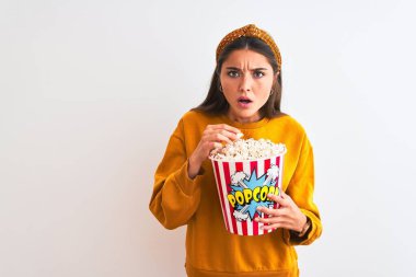 Genç ve güzel bir kadın, izole edilmiş beyaz arka planda duran patlamış mısırları tutuyor. Şaşırmış bir yüzle korkmuş ve korku ifadesinden heyecanlanmış.