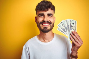 Dövmeli genç adam, izole edilmiş sarı arka planda dolarlar tutuyor mutlu bir yüzü var, gülümsüyor ve kendine güvenen bir gülümsemeyle dişlerini gösteriyor.
