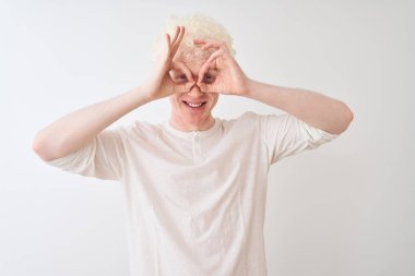 Genç albino sarışın adam, günlük tişört giyip izole edilmiş beyaz arka planda dikiliyor. Dürbün gibi jest yapıyor, gözleri parmaklarından bakıyor. Çılgın ifade.