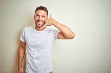 Genç yakışıklı adam giyiyor rahat beyaz tişört yapıyor gülümseyen izole arka plan üzerinde telefon hareketi ile el ve parmak telefonda konuşuyor gibi. Kavramları iletişim.