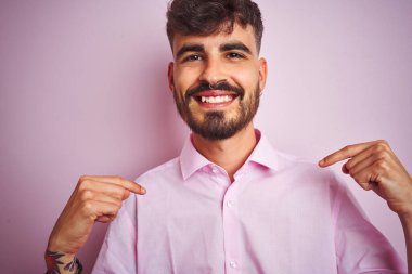 Dövme ile genç adam gömlek ayakta izole pembe arka plan üzerinde yüz gülümseme ile kendine güvenen görünümlü, parmaklarıyla gurur ve mutlu kendini işaret.