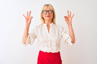 Tişört ve gözlük takan orta yaşlı bir iş kadını izole edilmiş beyaz arka planda durup rahatlıyor ve parmaklarıyla meditasyon hareketi yaparken gözleri kapalı gülümsüyor. Yoga konsepti.