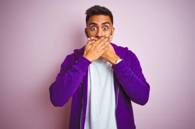 Mor eşofman üstü giyen genç Hintli adam izole edilmiş pembe arka planda dikiliyor. Ağzını yanlışlıkla ellerle kapatıyor. Gizli konsept.