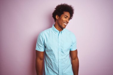 Afro saçlı, mavi gömlekli genç bir adam izole edilmiş pembe arka planda dikiliyor yüzünde gülümseme, doğal bir ifade ile yan yana bakıyor. Kendinden emin gülen.