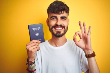 Alman pasaportlu genç adam izole edilmiş sarı arka plan üzerinde tamam işareti yapıyor, mükemmel sembol.