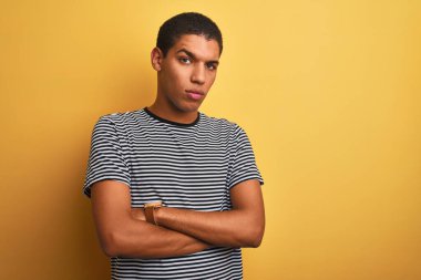 Genç yakışıklı Arap adam, izole edilmiş sarı arka plan şüphecisi ve gergin kolları çaprazlanmış yüz ifadesini tasvip etmeyen donanma tişörtü giyiyor. Negatif kişi.