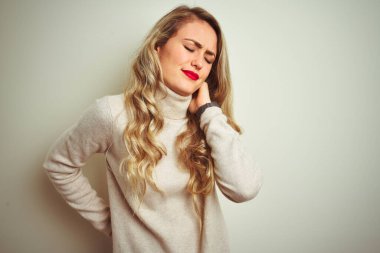 Güzel bir kadın kışın balıkçı yaka kazak giyiyor, beyaz arka planda boynu ağrıyor, boynuna elle dokunuyor, kas ağrısı çekiyor.