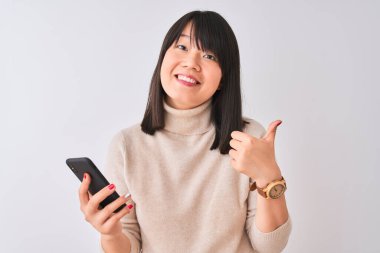 Genç, güzel Çinli bir kadın, izole edilmiş beyaz arka planda duran akıllı telefonu kullanıyor. Kocaman gülümsüyor ve gülümsüyor. İşaret veriyor.
