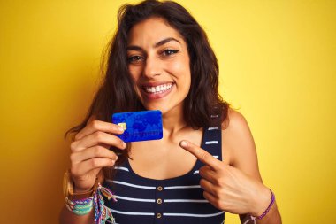 Sarı arka planda duran kredi kartı tutan genç ve güzel bir kadın. El ve parmak ile işaret ederek çok mutlu.