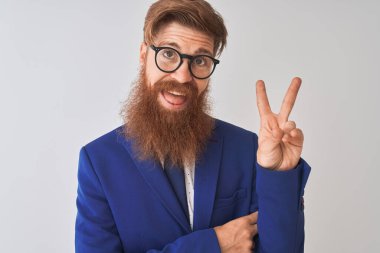 Genç, kızıl saçlı, takım elbiseli ve gözlüklü bir iş adamı izole edilmiş beyaz arka planda gülümsüyor ve kameraya gülümsüyor zafer işareti yapıyor. İki numara..