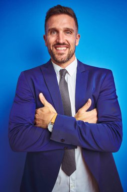 Mavi izole edilmiş arka planda, kollarını kavuşturup kameraya gülümseyen genç yakışıklı iş adamının dikey açısı. Pozitif kişi.