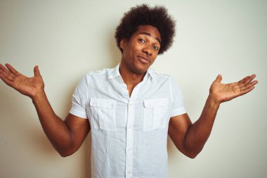 Afro saçlı, beyaz arka plan üzerinde duran, elleri ve kolları havada, kafası karışmış Afro-Amerikan bir adam. Şüphe kavramı.