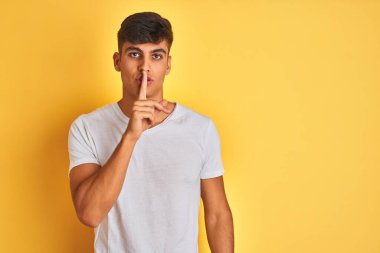 İzole edilmiş sarı arka planda duran beyaz tişörtlü genç Hintli adam sessiz olmak istiyor. Sessizlik ve gizli konsept.