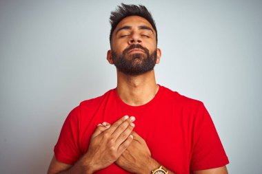 İzole edilmiş beyaz arka plan üzerinde kırmızı tişört giyen genç Hintli adam elleri göğsünde, gözleri kapalı ve yüzünde minnet dolu bir jest ile gülümsüyor. Sağlık kavramı.