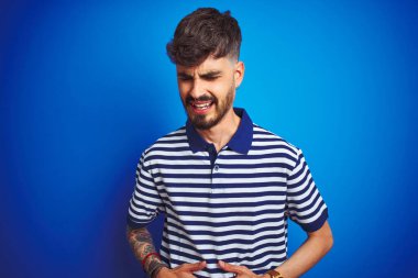 Çizgili polo giyen genç bir adam midesi ağrıyan, hazımsızlık çeken, acı veren bir hastalık yüzünden mavi arka planda duruyor. Ache kavramı.