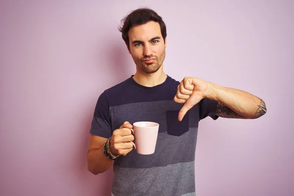 Young handsome man with tattoo drinking cup of coffee over isolated pink background with angry face, negative sign showing dislike with thumbs down, rejection concept