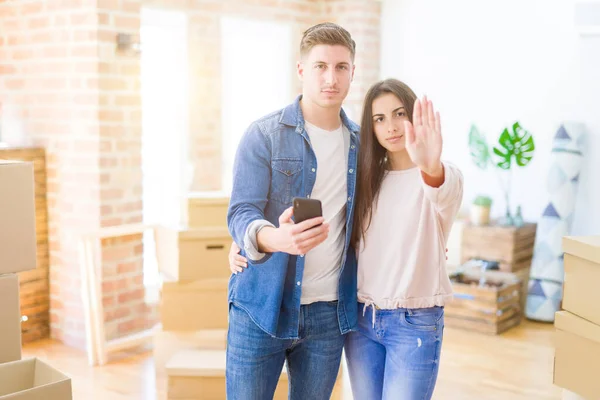 Beautiful young couple moving to a new house using smartphone with open hand doing stop sign with serious and confident expression, defense gesture