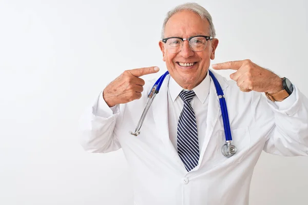 孤立した白い背景の上に立って聴診器を身に着けているシニア白髪の医者の男は 陽気に見せ 指の歯と口で指して笑っています 歯科衛生の概念 — ストック写真