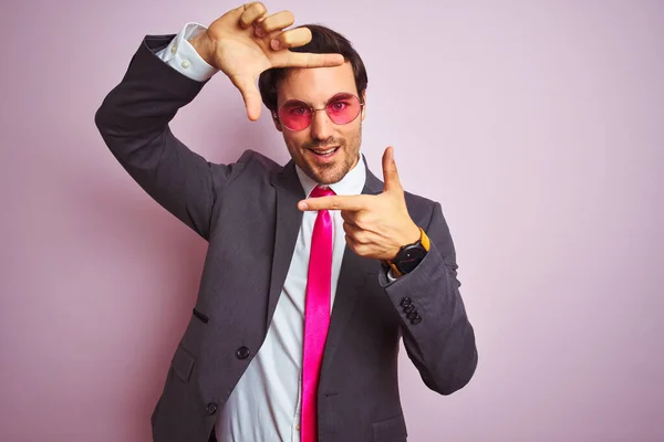 孤立したピンクの背景にスーツとサングラスを身に着けている若いハンサムなビジネスマンは 幸せな顔で手と指でフレームを作り笑顔 創造性と写真の概念 — ストック写真
