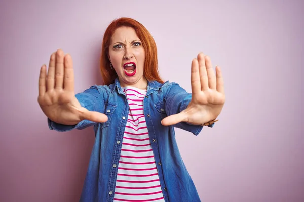Beautiful redhead woman wearing denim shirt and striped t-shirt over isolated pink background doing stop gesture with hands palms, angry and frustration expression