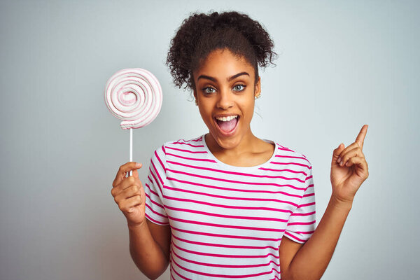 Африканская американская подросток женщина ест красочные конфеты на изолированном белом фоне очень счастливы указывая рукой и пальцем в сторону
