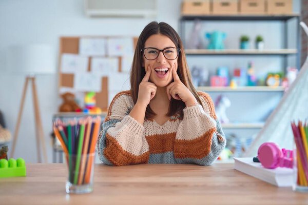 Young beautiful teacher woman wearing sweater and glasses sitting on desk at kindergarten Smiling with open mouth, fingers pointing and forcing cheerful smile