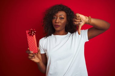Genç Afrikalı Amerikalı bir kadın sevgililer günü hediyesini elinde tutuyor. Kızgın yüzüyle izole edilmiş kırmızı arka planda duruyor. Negatif işaretler baş parmakları aşağıda, reddedilme kavramından hoşlanmadığını gösteriyor.