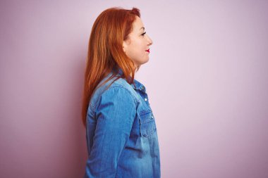 Güzel kızıl saçlı kadın kot gömlek ve çizgili t-shirt giyiyor. İzole edilmiş pembe arka plan üzerinde. Yan tarafa bakıyor, rahat profil pozu veriyor ve kendine güvenen bir gülümseme takınıyor..