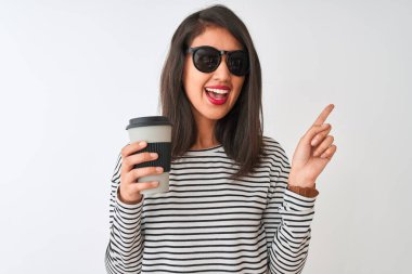 Güneş gözlüğü takan Çinli bir kadın izole edilmiş beyaz arka planda kahve içerken mutlu bir şekilde elini ve parmağını kenarı işaret ediyor.