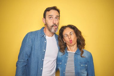Güzel bir orta yaşlı çift, izole edilmiş sarı arka planda kot gömlek giyip dudaklı balık suratlı, çılgın ve komik bir jest yapıyorlar. Komik ifade.