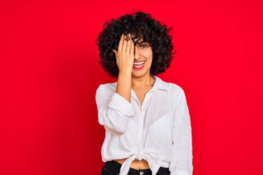 Kıvırcık saçlı, beyaz tişörtlü, izole edilmiş kırmızı arka planı olan genç bir Arap kadın bir gözünü eliyle kapatıyor, yüzünde kendine güvenen bir gülümseme ve şaşırtıcı bir duygu var..