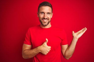 Kırmızı izole edilmiş arka plan üzerine gündelik tişört giyen genç yakışıklı adam avuç içini gösteriyor ve baş parmaklarıyla hareket ediyor, mutlu ve neşeli gülümsüyor.