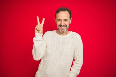 Yakışıklı, orta yaşlı, gri saçlı, izole edilmiş kırmızı arka planda gülümseyen ve parmaklarını zafer işareti yaparken gösteren bir adam. İki numara..