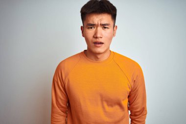 Turuncu süveter giyen Asyalı genç adam beyaz arka planda duruyor. Şaşırmış ve korkmuş bir ifade, korku ve heyecanlı bir yüz ifadesi var..