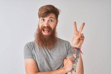 İzole edilmiş gri arka planda dikilen kızıl saçlı genç adam zafer işareti yaparken kameraya gülümsüyor. İki numara..