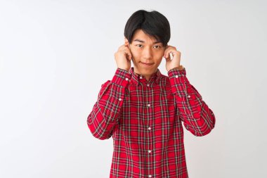 İzole edilmiş beyaz arka planda dikilen gündelik kırmızı tişörtlü genç Çinli adam yüksek sesli müzik sesi için rahatsız edici ifadeleri olan parmaklarıyla kulakları kapatıyor. Sağır kavramı.