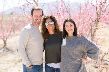 Üç gülümseyen güzel ailesi neşeli ve sarılma şeftali bahçesinde bahar güneşli gün zevk pembe yaprakları ile