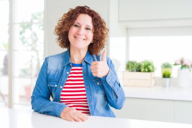 Kıvırcık saçlı, kot ceket giyen orta yaşlı bir kadın evde el hareketleriyle mutlu parmaklar yapıyor. Kameraya bakıp başarıyı gösteren ifade onaylanıyor.