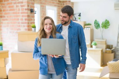 Yeni bir daireye taşımak için mutlu karton kutular etrafında bir odada bilgisayar laptop ayakta kullanarak genç çift