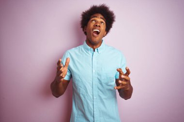 Afro saçlı, mavi gömlekli genç bir adam pembe arka planda dikiliyor deli deli deli bağırıyor ve agresif bir ifade ve kollar havada bağırıyor. Hayal kırıklığı konsepti.