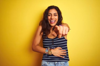 İzole edilmiş sarı arka planda duran çizgili tişört giyen genç ve güzel bir kadın sana gülüyor, kamerayı işaret ediyor, vücudunu gösteriyor, utanç ifadesi var.