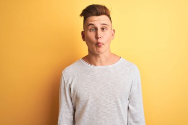 İzole edilmiş sarı arka plan üzerinde duran beyaz tişörtlü genç yakışıklı adam dudaklı balık suratı yapıyor, çılgınca ve komik bir jest. Komik ifade.