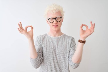İzole edilmiş beyaz arka plan üzerine çizgili tişört ve gözlük takan genç albino sarışın adam rahatla ve gözleri kapalı meditasyon yaparken parmaklarıyla gülümse. Yoga konsepti.
