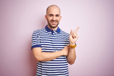 Sakallı genç ve kel bir adam pembe izole edilmiş arka planda günlük mavi tişört giyiyor. Yüzünde kocaman bir gülümseme var. Elleri ve parmakları kameraya bakıyor..