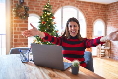 Güzel bir kadın evde dizüstü bilgisayarla çalışıyor Noel ağacının etrafında kameraya bakıyor kucaklaşmak için kollarını açmış gülümsüyor. Mutluluğu kucaklayan neşeli bir ifade..