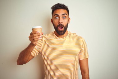 Genç Hintli bir adam, izole edilmiş beyaz arka planda duran bir fincan kahve içiyor. Şaşırmış bir yüz ifadesinden korkuyor ve heyecanlanıyor.