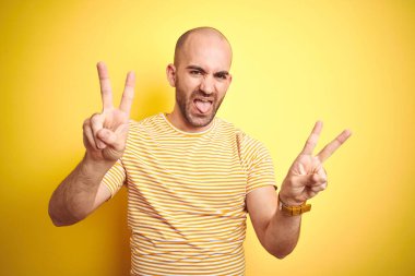 Sakallı, sarı izole edilmiş arka plan üzerine günlük çizgili tişört giyen genç ve kel bir adam iki elini de zafer işareti yaparken gösteren bir dille gülüyor. İki numara..