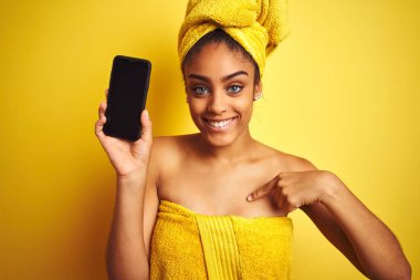 Duş üstüne havlu giyen Afro kadın izole edilmiş sarı arka plan üzerinde smatrphone gösteriyor.